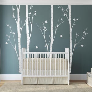 May include: A white wall decal featuring a forest of birch trees with slender trunks and branches, accented by small flying birds. The decal is applied to a dark teal wall behind a white wooden crib with a beige mattress and skirt.