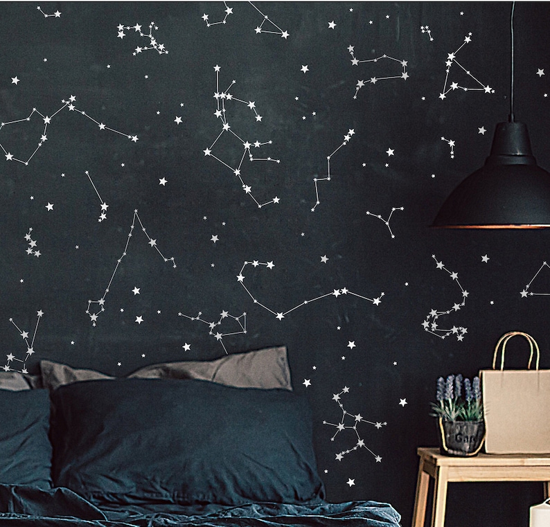 Puede incluir: Una cama azul oscuro con calcoman&iacute;as de pared de constelaciones de estrellas blancas. La cama tiene dos almohadas y una manta azul oscuro. Una l&aacute;mpara negra cuelga del techo sobre la cama. Una peque&ntilde;a mesa de madera con una planta en maceta y una bolsa de papel marr&oacute;n se encuentra en la esquina de la habitaci&oacute;n.