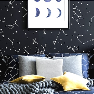 Puede incluir: Un dormitorio blanco y negro con una cama que presenta una cabecera azul marino y un edred&oacute;n con estampado gris y blanco. La pared est&aacute; decorada con constelaciones y estrellas blancas. Hay varias almohadas en la cama, incluyendo dos almohadas amarillas, una almohada gris y una almohada blanca con un patr&oacute;n de rayas grises.