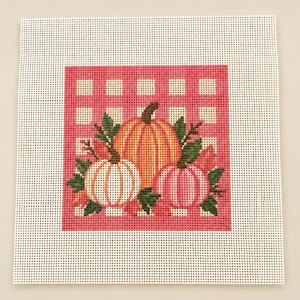 Könnte beinhalten: Ein quadratisches Stickleinen mit einem zentralen Motiv aus drei Kürbissen in Orange, Weiß und Rosa, umgeben von grünen Blättern. Die Kürbisse sind vor einem rosa-weiß karierten Vichy-Muster auf einem weißen Netzgewebe dargestellt.