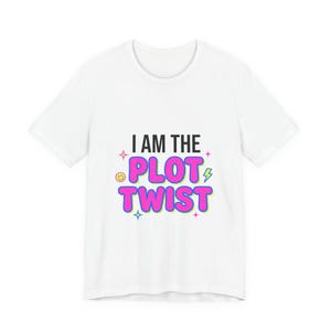Puede incluir: Una camiseta blanca de manga corta con el texto negro I AM THE y el texto rosa brillante PLOT TWIST. El diseño incluye pequeños elementos decorativos como una cara sonriente amarilla, un rayo verde y cuatro pequeñas estrellas rosas.