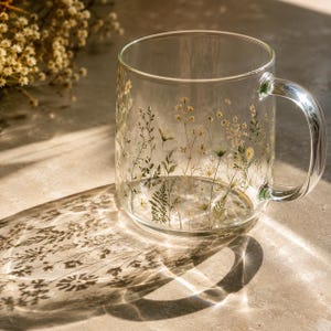 May include: A clear glass mug featuring a delicate botanical print of various wildflowers, grasses, and ferns in shades of green, yellow, and white. The mug has a smooth, cylindrical body with a curved handle and casts a detailed floral shadow.
