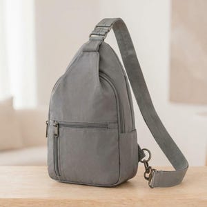 May include: A grey sling backpack featuring a teardrop shape, a single adjustable shoulder strap, and a front zippered pocket. The bag is constructed from a smooth, solid-colored fabric and includes metal hardware, such as zippers and a D-ring attachment.
