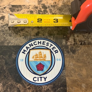 Può includere: Una toppa circolare con il logo del Manchester City, con un diametro di circa 7,6 cm. Il logo include uno scudo con una nave, una rosa rossa e la scritta "MANCHESTER CITY". È visibile un metro a nastro.