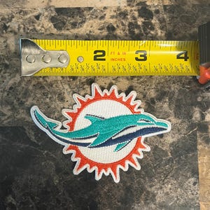 May include: Embroidered patch featuring the Miami Dolphins logo. The patch includes a teal dolphin leaping through a sunburst design with orange and white accents. The patch measures approximately 3 inches in length.
