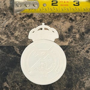 Puede incluir: Un emblema blanco del Real Madrid con una corona, de aproximadamente 6,35 cm de di&aacute;metro. Una cinta m&eacute;trica amarilla se coloca encima del emblema. El emblema est&aacute; sobre una superficie oscura y texturizada.