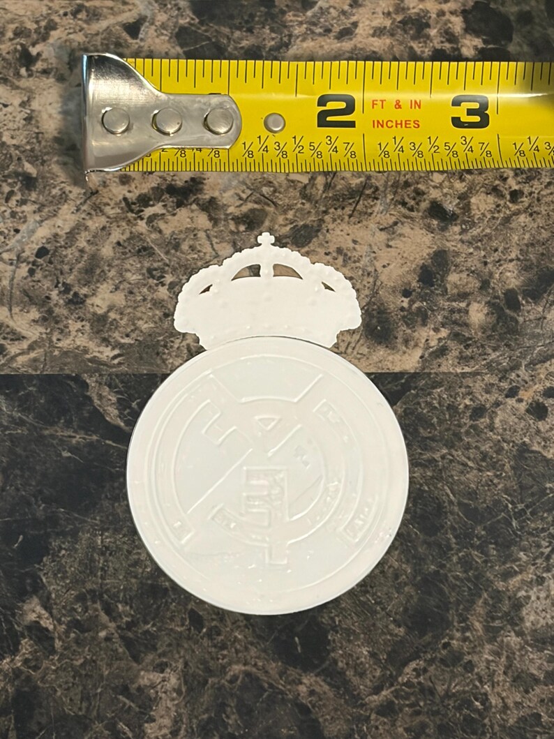 Puede incluir: Un emblema blanco del Real Madrid con una corona, de aproximadamente 6,35 cm de di&aacute;metro. Una cinta m&eacute;trica amarilla se coloca encima del emblema. El emblema est&aacute; sobre una superficie oscura y texturizada.