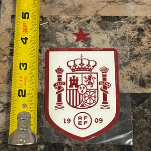 Puede incluir: Un parche rojo y blanco en forma de escudo con el emblema de la RFEF, una corona, columnas y el texto PLVS VLTRA y 19 09. Una estrella roja se sitúa sobre el escudo. Una cinta métrica amarilla muestra una altura de unos 11,4 cm.