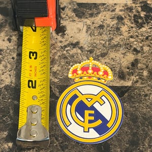 Puede incluir: Un emblema del Real Madrid, con una corona y un dise&ntilde;o circular con las letras "RMF", se muestra junto a una cinta m&eacute;trica. El emblema tiene aproximadamente 6,35 cm de di&aacute;metro. Los colores son amarillo, azul, blanco y rojo.