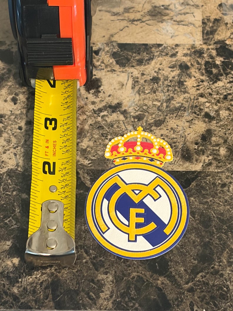 Puede incluir: Un emblema del Real Madrid, con una corona y un dise&ntilde;o circular con las letras "RMF", se muestra junto a una cinta m&eacute;trica. El emblema tiene aproximadamente 6,35 cm de di&aacute;metro. Los colores son amarillo, azul, blanco y rojo.