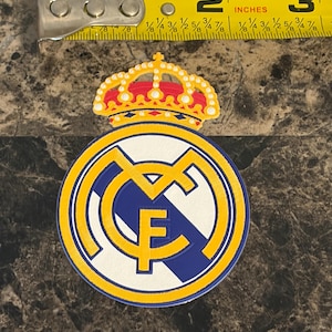 Puede incluir: Parche circular con el escudo del Real Madrid. El dise&ntilde;o presenta un fondo blanco con detalles azules y amarillos. El escudo incluye una corona y las letras "RMF". El parche mide aproximadamente 5 cm de di&aacute;metro.