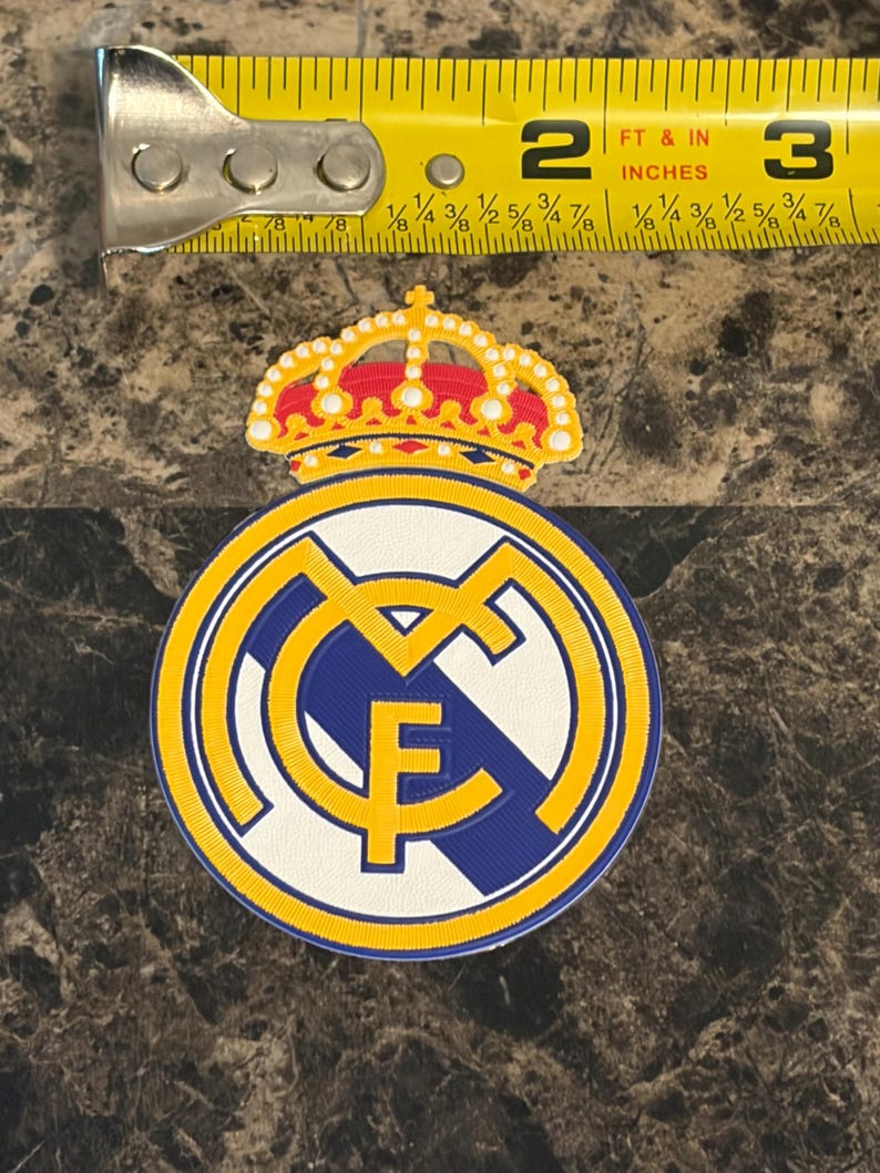 Puede incluir: Parche circular con el escudo del Real Madrid. El dise&ntilde;o presenta un fondo blanco con detalles azules y amarillos. El escudo incluye una corona y las letras "RMF". El parche mide aproximadamente 5 cm de di&aacute;metro.
