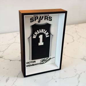 Pode incluir: Uma caixa expositora retangular com uma camisola de basquetebol preta dos San Antonio Spurs com o nome Wembanyama e o número 1 em branco. Acima da camisola está o logótipo dos Spurs e abaixo o nome Victor Wembanyama com uma assinatura.