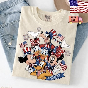 May include: An ivory t-shirt featuring a graphic of Disney characters wearing patriotic hats and holding American flags. The design includes fireworks and a blue banner with the text USA ANNIVERSARY 1776-2026. The shirt has a Comfort Colors label.