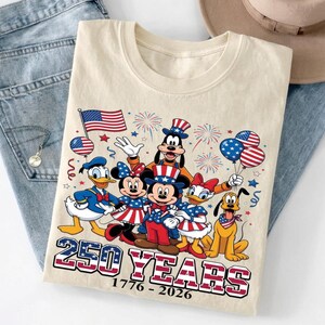 Może przedstawiać: Kremowy t-shirt z grafiką przedstawiającą postacie Disneya, w tym Myszkę Miki, Minnie, Kaczora Donalda, Daisy, Goofy'ego i Pluto w patriotycznych strojach. Projekt zawiera amerykańską flagę, fajerwerki oraz napis 250 YEARS 1776-2026.