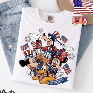 May include: A white t-shirt featuring a graphic of Mickey Mouse, Minnie Mouse, Donald Duck, Daisy Duck, Goofy, and Pluto wearing patriotic hats. The design includes American flags, fireworks, and a banner reading USA 250TH ANNIVERSARY 1776-2026.