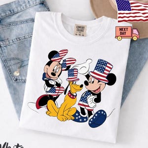 May include: A white t-shirt featuring a graphic of Minnie Mouse, Pluto, and Mickey Mouse wearing patriotic top hats and clothing with American flag patterns. The shirt has a Comfort Colors label at the neck and is displayed on blue denim fabric.
