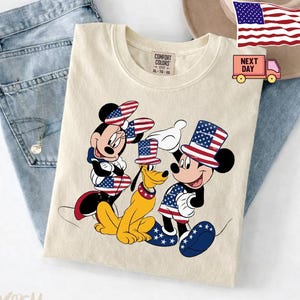 May include: An ivory t-shirt featuring a graphic of Minnie Mouse, Pluto, and Mickey Mouse wearing patriotic American flag-themed hats and clothing. The shirt has a Comfort Colors label at the neck and is displayed alongside blue denim jeans.