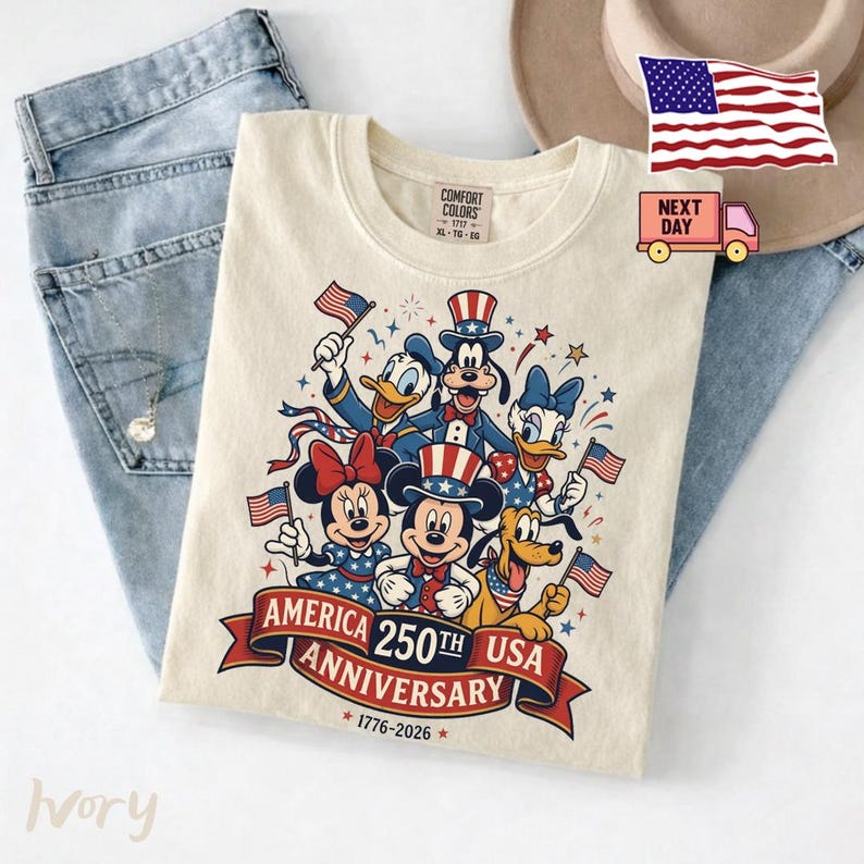 May include: An ivory t-shirt featuring a graphic of six cartoon characters wearing patriotic hats and holding American flags. Below the characters, a red banner displays the text AMERICA 250TH USA ANNIVERSARY 1776-2026 in bold lettering.