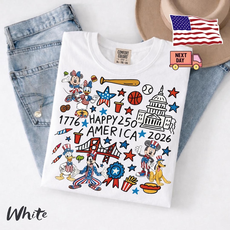 May include: A white t-shirt featuring a colorful graphic print with cartoon characters, the US Capitol building, the Golden Gate Bridge, stars, and food items. The text reads 1776 HAPPY 250 AMERICA 2026. The shirt is layered over blue denim jeans.