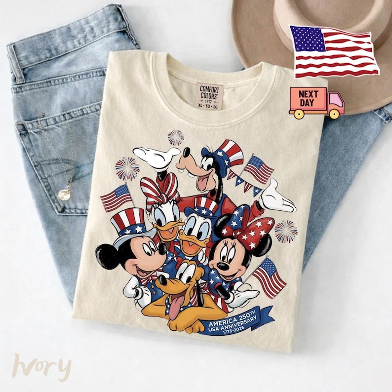 May include: An ivory t-shirt featuring a graphic of Disney characters wearing patriotic hats and holding American flags. The design includes fireworks and a blue banner with the text USA ANNIVERSARY 1776-2026. The shirt has a Comfort Colors label.