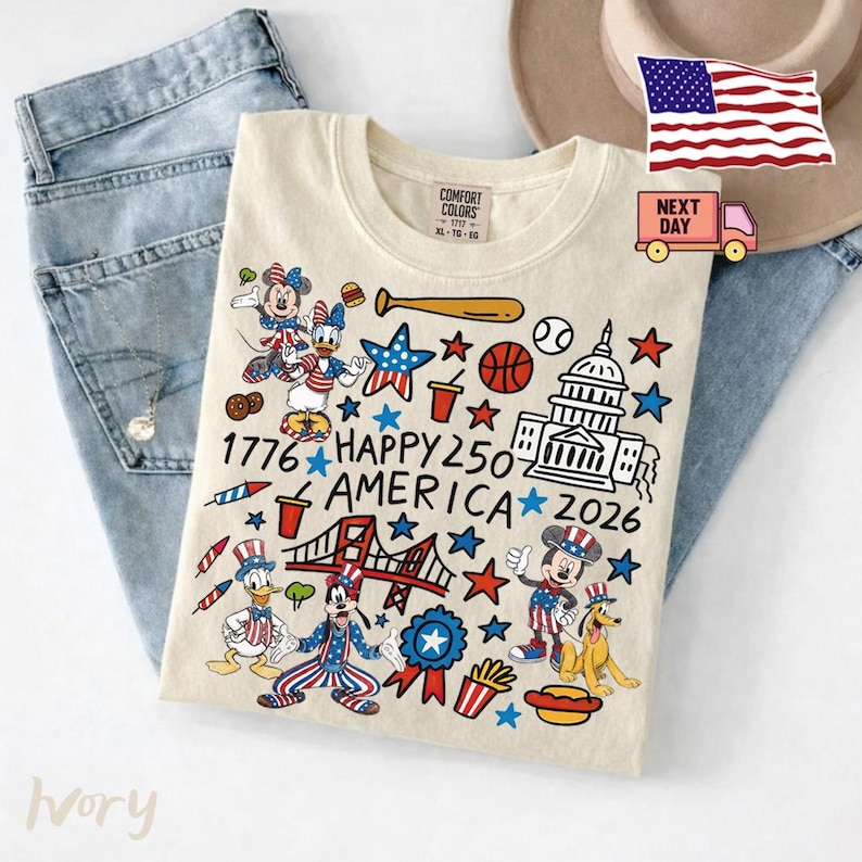 May include: An ivory t-shirt featuring a graphic print with the text 1776 HAPPY 250 AMERICA 2026. The design includes cartoon characters, the U.S. Capitol building, a bridge, a baseball bat, a basketball, stars, and food items in red, white, and blue.