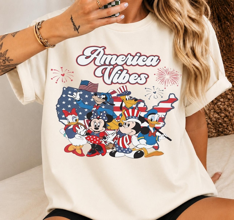 May include: A cream-colored t-shirt featuring a graphic print with the text "America Vibes" in red and white. The design includes a map of the United States filled with a flag pattern, fireworks, and several cartoon characters wearing patriotic outfits.