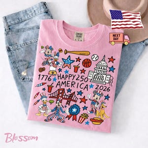 May include: A light pink t-shirt featuring a graphic print with the text 1776 HAPPY 250 AMERICA 2026. The design includes cartoon characters wearing patriotic outfits, the U.S. Capitol building, a bridge, sports equipment, food items, and stars.