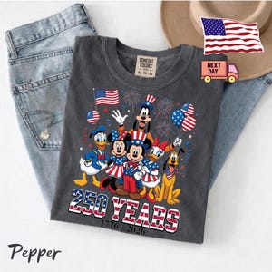 K&ouml;nnte beinhalten: Ein anthrazitfarbenes T-Shirt mit einer Grafik von Disney-Figuren wie Mickey Maus, Minnie Maus, Donald Duck, Daisy Duck, Goofy und Pluto in patriotischer Kleidung. Der Text lautet 250 YEARS 1776-2026, erg&auml;nzt durch Feuerwerk.