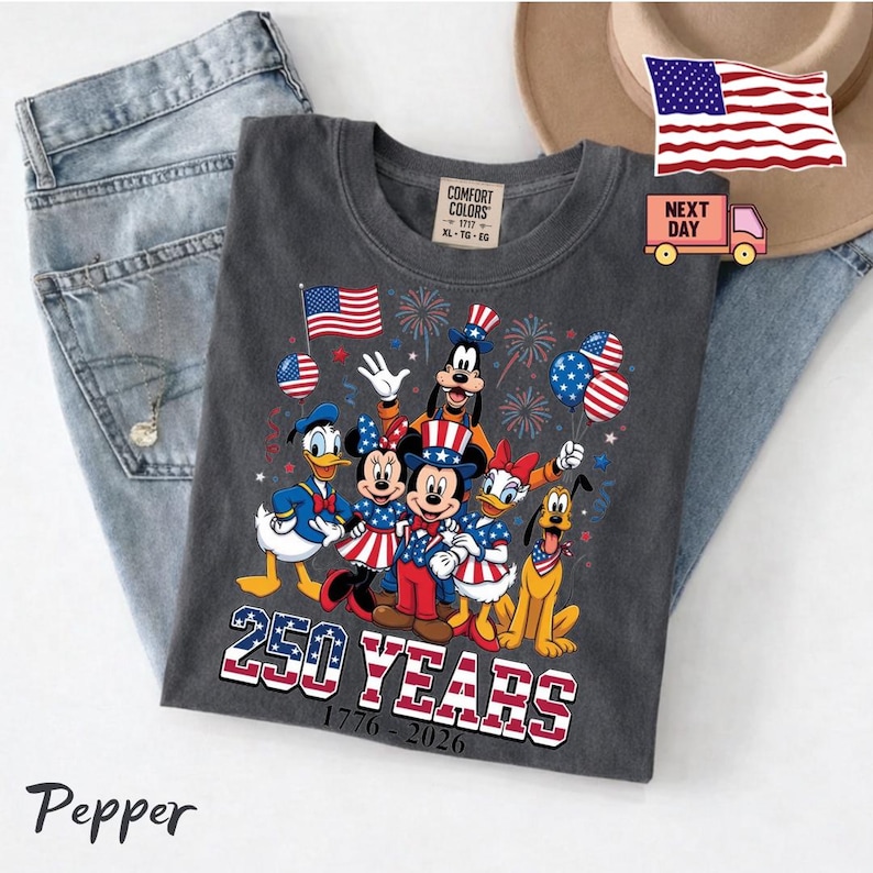 K&ouml;nnte beinhalten: Ein anthrazitfarbenes T-Shirt mit einer Grafik von Disney-Figuren wie Mickey Maus, Minnie Maus, Donald Duck, Daisy Duck, Goofy und Pluto in patriotischer Kleidung. Der Text lautet 250 YEARS 1776-2026, erg&auml;nzt durch Feuerwerk.