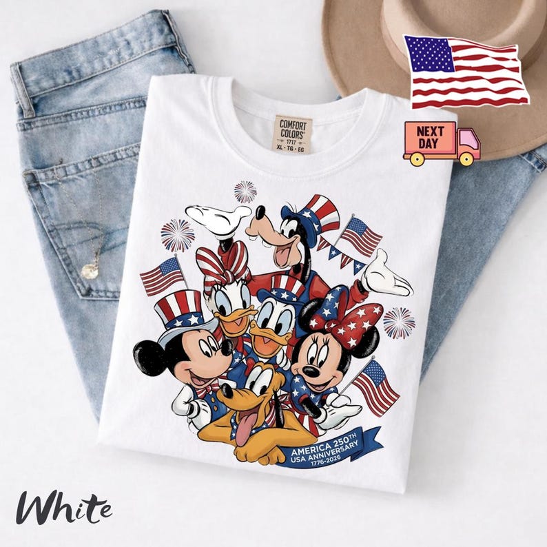 May include: A white t-shirt featuring a graphic of Mickey Mouse, Minnie Mouse, Donald Duck, Daisy Duck, Goofy, and Pluto wearing patriotic hats and bows. The design includes American flags, fireworks, and text reading America 250th USA Anniversary 1776-2026.