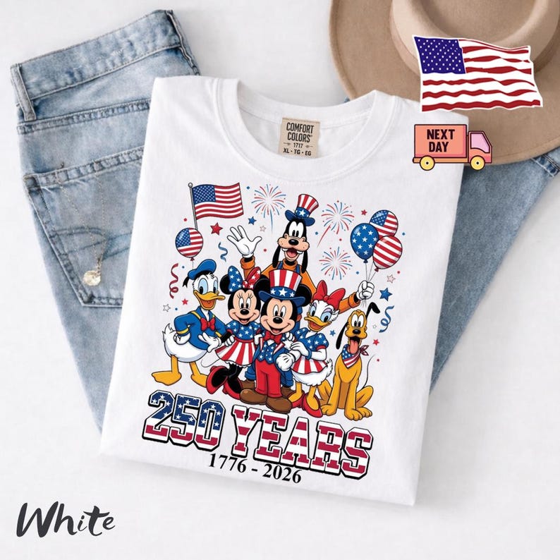 K&ouml;nnte beinhalten: Ein wei&szlig;es T-Shirt mit einer Grafik von Zeichentrickfiguren in patriotischen rot-wei&szlig;-blauen Outfits. Das Design umfasst Feuerwerk, Luftballons und den Text 250 YEARS 1776-2026. Das Shirt hat ein Comfort Colors Etikett am inneren Halsausschnitt.