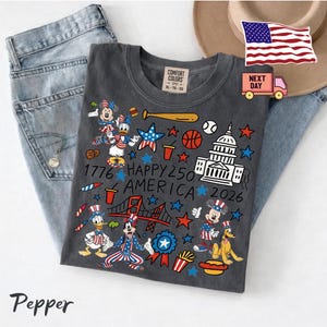 May include: A charcoal gray t-shirt featuring a patriotic graphic print with cartoon characters, the US Capitol building, a bridge, sports equipment, and the text 1776 HAPPY 250 AMERICA 2026. The shirt has a Comfort Colors label at the inner neckline.
