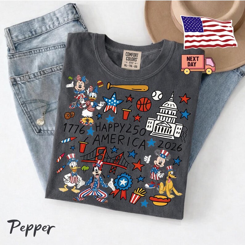 May include: A charcoal gray t-shirt featuring a patriotic graphic print with cartoon characters, the US Capitol building, a bridge, sports equipment, and the text 1776 HAPPY 250 AMERICA 2026. The shirt has a Comfort Colors label at the inner neckline.