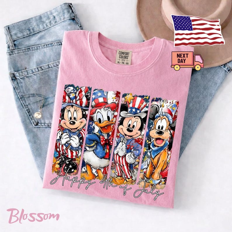 May include: A light pink t-shirt featuring a graphic print of four cartoon characters wearing patriotic red, white, and blue hats and outfits. The text "Happy 4th of July" is printed in a cursive script below the characters. The shirt has a Comfort Colors label.