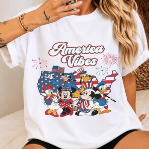 May include: A white t-shirt featuring a graphic print with the text America Vibes in a retro font. The design includes a map of the United States filled with stars and stripes, surrounded by fireworks and cartoon characters wearing patriotic outfits.