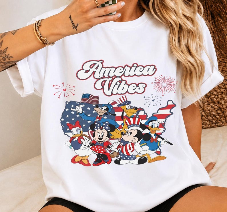May include: A white t-shirt featuring a graphic print with the text America Vibes in a retro font. The design includes a map of the United States filled with stars and stripes, surrounded by fireworks and cartoon characters wearing patriotic outfits.