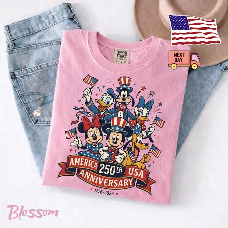 May include: A light pink t-shirt featuring a graphic of Mickey Mouse, Minnie Mouse, Donald Duck, Daisy Duck, Goofy, and Pluto wearing patriotic hats. The text reads AMERICA 250TH ANNIVERSARY USA 1776-2026. The shirt is displayed with blue jeans.