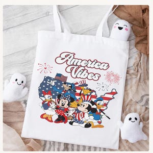 May include: A white tote bag featuring a graphic print of the text America Vibes above a map of the United States filled with a flag pattern. The design includes cartoon characters wearing patriotic hats and clothing, surrounded by red firework icons.