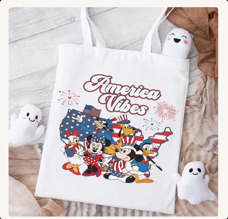 May include: A white tote bag featuring a graphic print of the text America Vibes above a map of the United States filled with a flag pattern. The design includes cartoon characters wearing patriotic hats and clothing, surrounded by red firework icons.