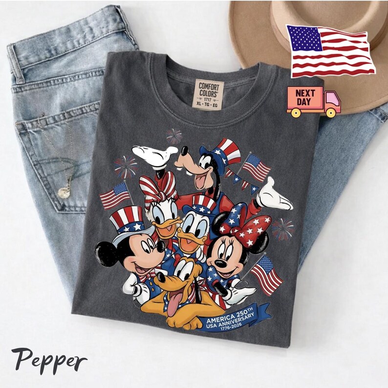 May include: A charcoal grey t-shirt featuring a graphic of cartoon characters wearing patriotic hats and outfits, surrounded by American flags and fireworks. The text reads America 250th Anniversary 1776-2026. The shirt has a Comfort Colors brand label.