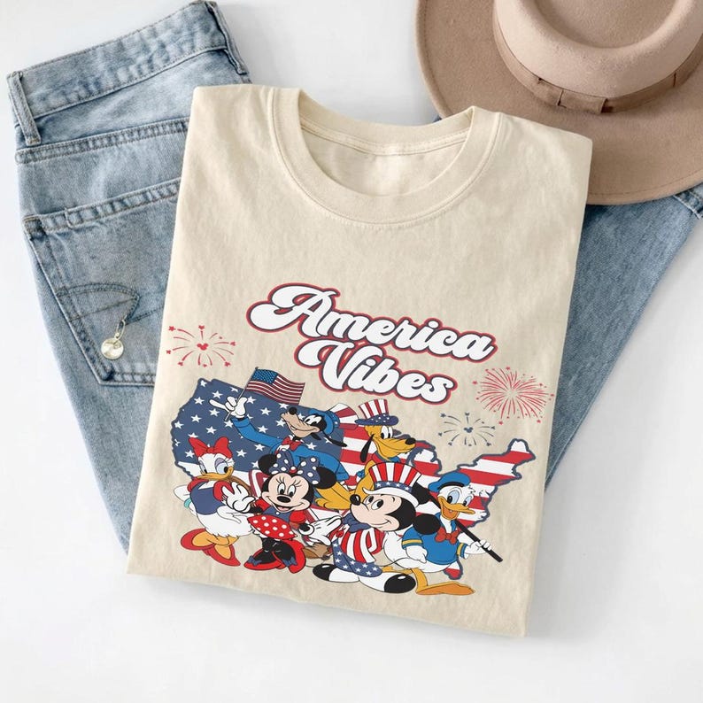 May include: A cream-colored t-shirt featuring the text America Vibes in red and white script. The graphic includes a map of the United States filled with stars and stripes, surrounded by cartoon characters wearing patriotic hats and fireworks.