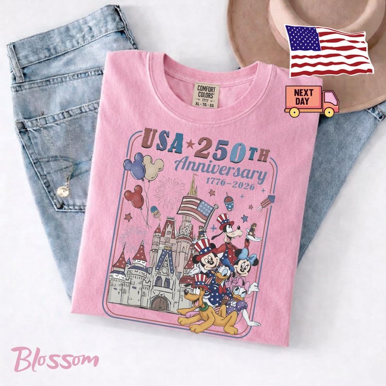 Peut inclure: Un t-shirt rose clair avec un motif repr&eacute;sentant un ch&acirc;teau, des feux d'artifice et des personnages de dessin anim&eacute; portant des chapeaux patriotiques. Le texte indique USA 250TH Anniversary 1776-2026. Le t-shirt est pos&eacute; sur un jean bleu et un chapeau beige.
