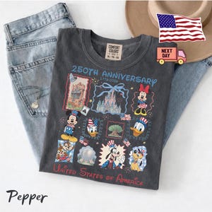 May include: A charcoal grey t-shirt featuring a 250th Anniversary 1776-2026 graphic with Disney characters, landmarks, and the text United States of America. The shirt has a Comfort Colors label, and the design includes Mickey, Minnie, Donald, Goofy, Pluto, Chip, Dale, and patriotic motifs.