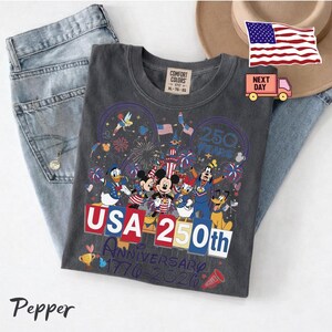 May include: A charcoal gray t-shirt featuring a graphic of Disney characters including Mickey Mouse, Minnie Mouse, Donald Duck, Daisy Duck, Goofy, Pluto, and Tinker Bell. The text reads USA 250th Anniversary 1776-2026 with fireworks and a castle.