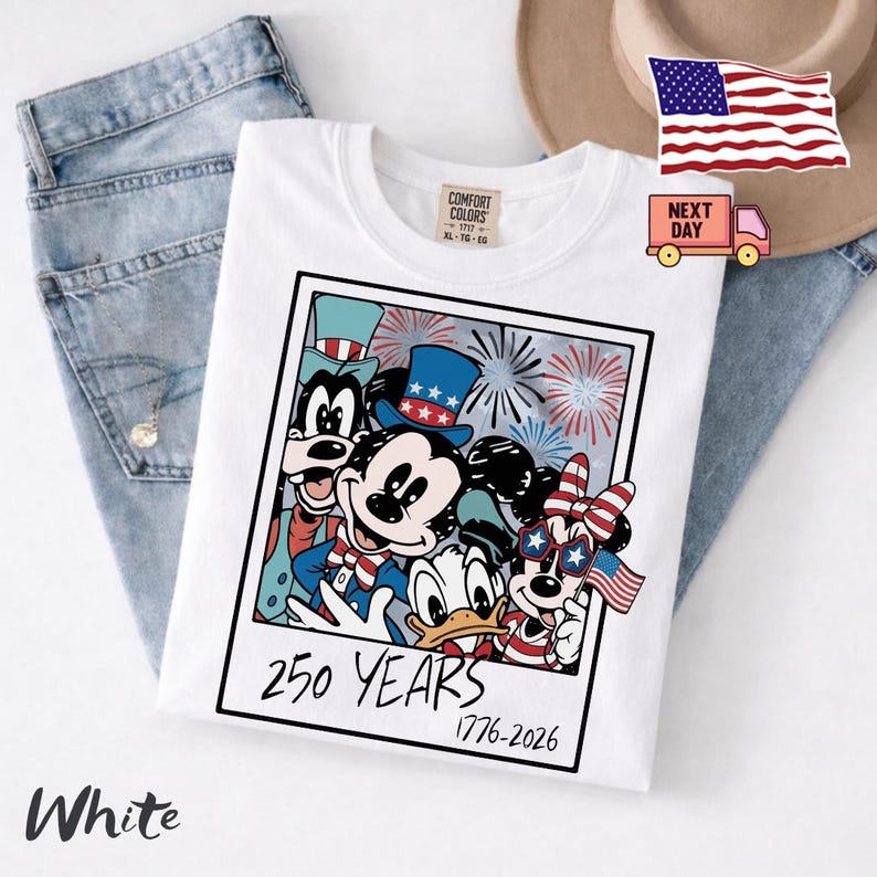 May include: A white t-shirt featuring a graphic print of cartoon characters wearing patriotic hats and accessories. The design is framed like a polaroid photo with the text 250 YEARS 1776-2026 printed at the bottom. The shirt has a Comfort Colors label.