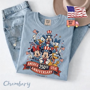 May include: A light blue t-shirt featuring a graphic of cartoon characters wearing patriotic hats and holding American flags. The text reads AMERICA 250TH USA ANNIVERSARY 1776-2026. The shirt has a Comfort Colors label at the neck and is paired with blue jeans.