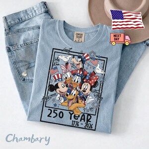 May include: A light blue t-shirt featuring a graphic of Disney characters including Mickey Mouse, Minnie Mouse, Donald Duck, Daisy Duck, Goofy, and Pluto wearing patriotic red, white, and blue attire. The text reads 250 Year 1776-2026.