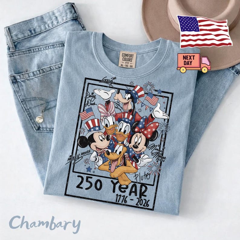 May include: A light blue t-shirt featuring a graphic of Disney characters including Mickey Mouse, Minnie Mouse, Donald Duck, Daisy Duck, Goofy, and Pluto wearing patriotic red, white, and blue attire. The text reads 250 Year 1776-2026.