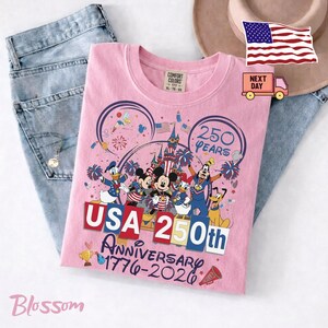Może przedstawiać: Jasnor&oacute;żowy t-shirt z grafiką przedstawiającą postacie Disneya, w tym Myszkę Miki, Kaczora Donalda i Goofy'ego. Wz&oacute;r zawiera tekst USA 250th Anniversary 1776-2026, zamek, fajerwerki i amerykańskie flagi. Koszulka posiada metkę Comfort Colors.
